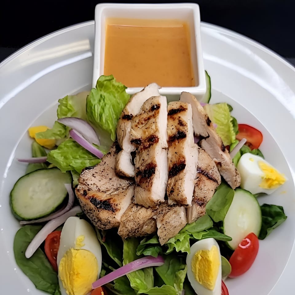 Grilled chicken salad with lettuce, cucumber, tomato, hard-boiled eggs, and a side of dressing.