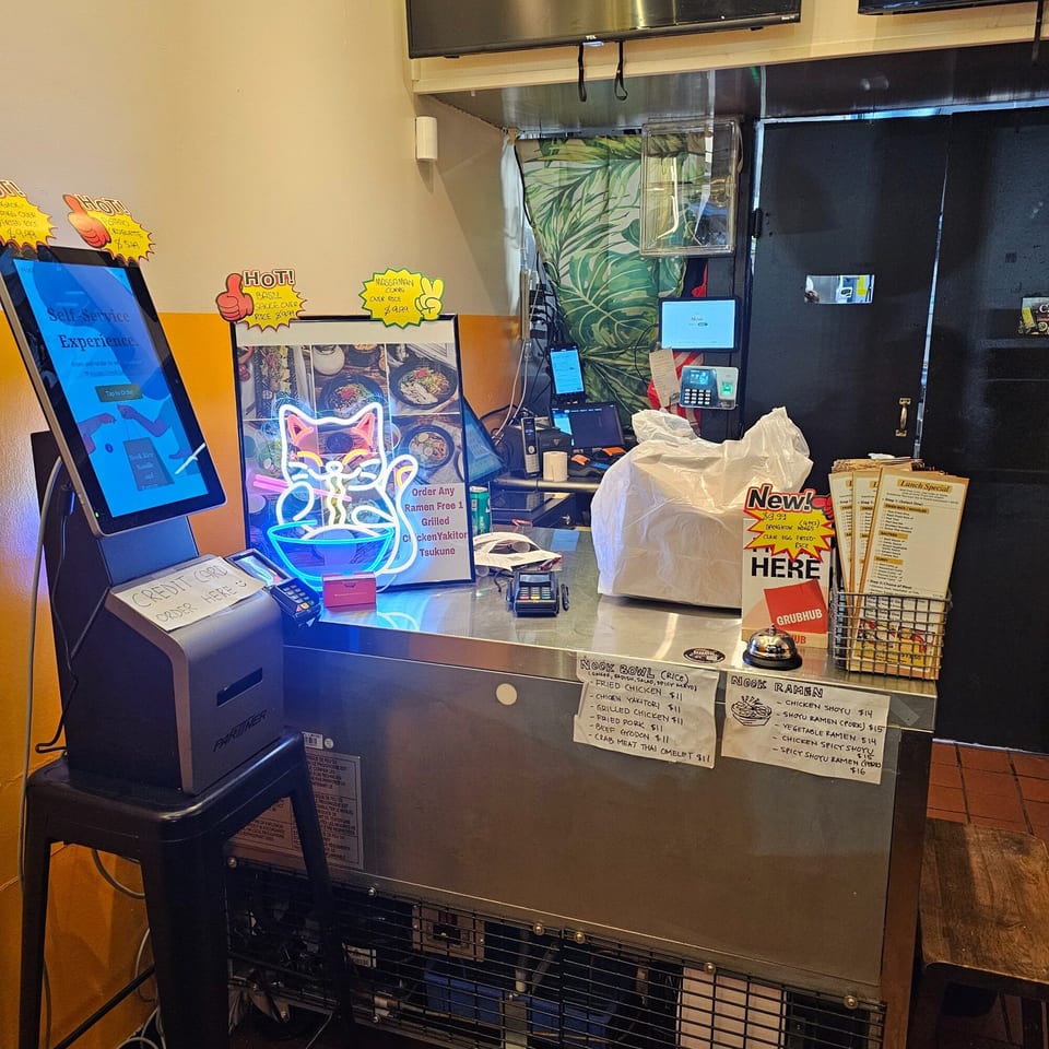A self-service ordering kiosk and counter with menus and a neon cat sign at Nook Thai restaurant.