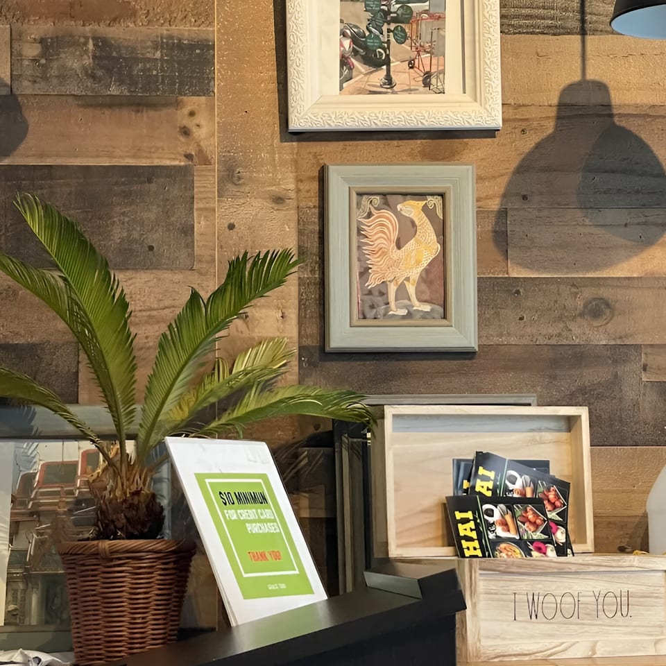 A Thai restaurant interior with a plant, framed art, and a sign on a wooden wall.
