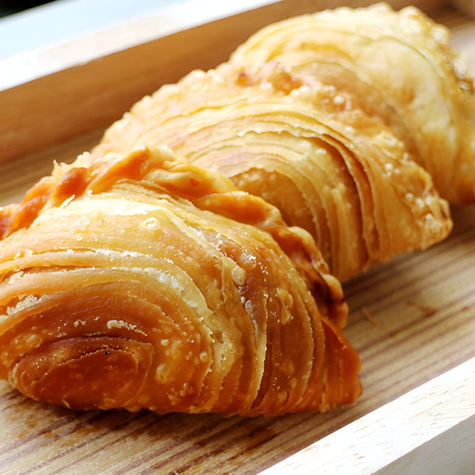 Three golden-brown, flaky Thai curry puffs with crimped edges, resting on a light wooden tray.